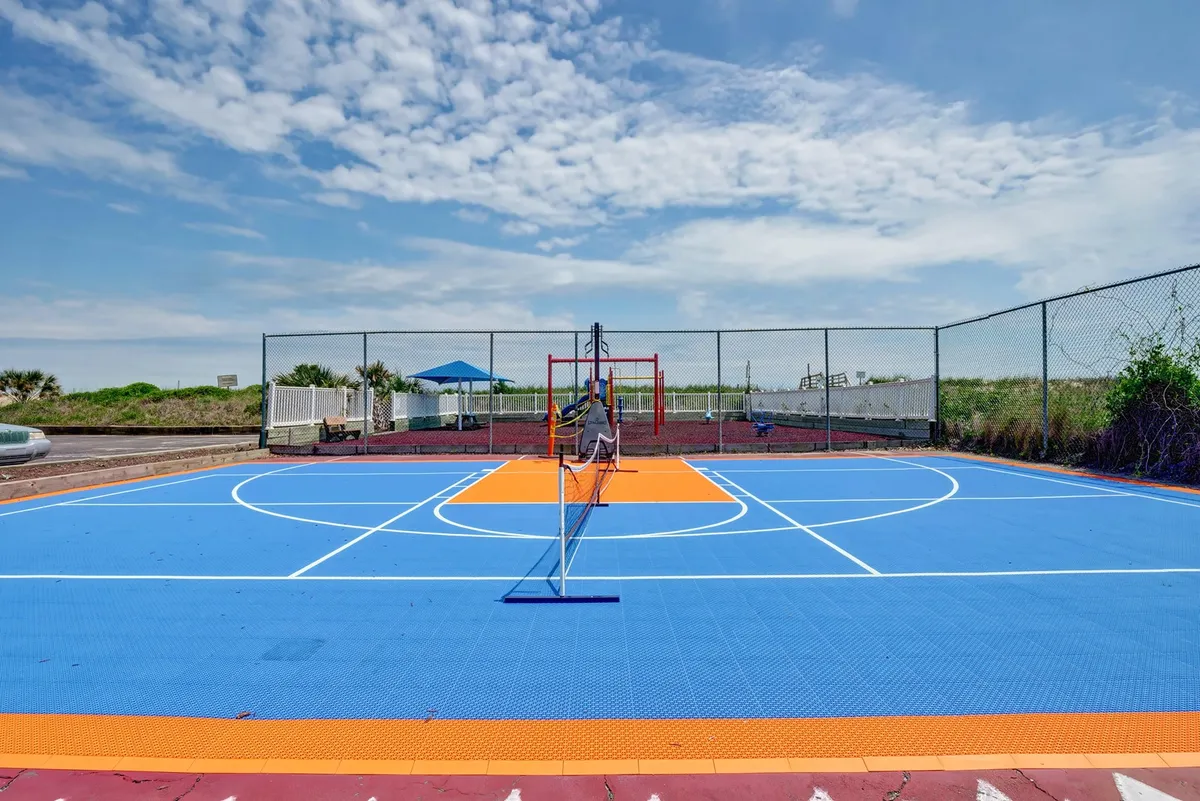 A vibrant exterior scene featuring a brightly colored basketball court with a tennis net, adjacent to modern playground equipment. The backdrop is a clear blue sky, and the area emanates an active, family-friendly atmosphere, with space for basketball and tennis games as well as children playing on the playground.