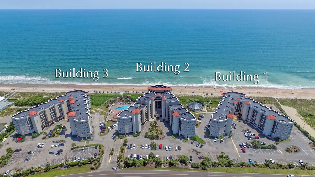 The resort's exterior is shown with three beachfront buildings, a sparkling swimming pool, and immediate access to the sandy beach. The area exudes a relaxed atmosphere, ideal for swimming, sunbathing, or a leisurely walk by the ocean at sunset.