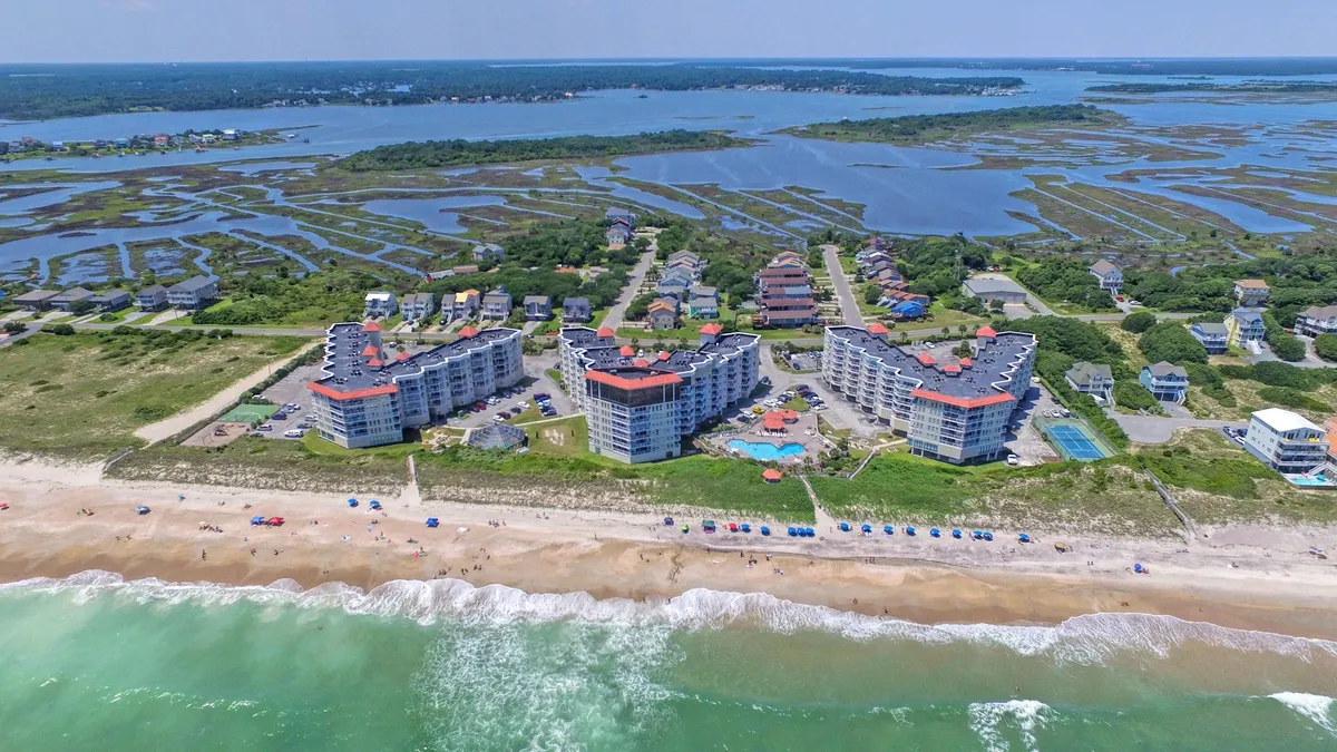 A sweeping exterior view of beachfront buildings with a large swimming pool, umbrellas on the sand, and lush greenery. The scene captures both the inviting pool and the beach just steps away, highlighting opportunities for sunbathing, swimming, poolside gatherings, and walks on the shore.