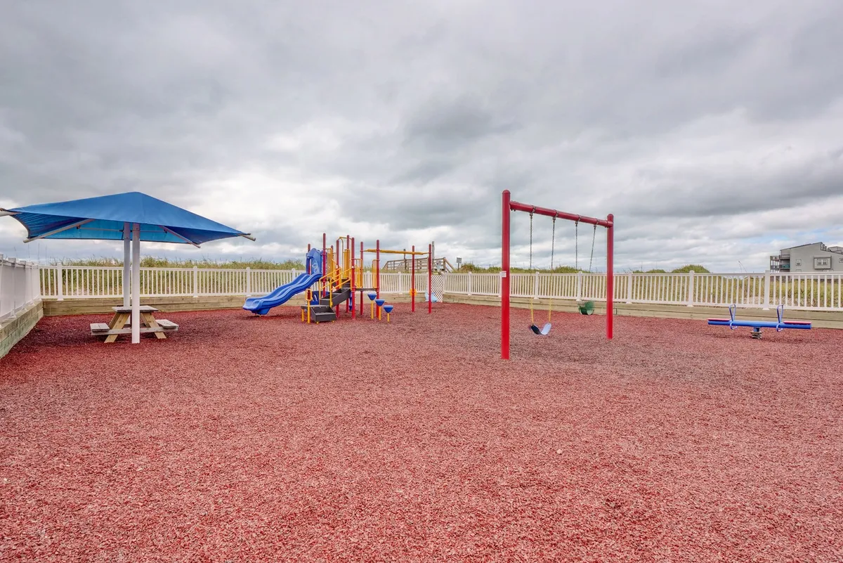 A cheerful outdoor playground with colorful equipment, swings, and a nearby picnic table. The setting shows children playing while adults can relax and enjoy a meal or snack at the table.