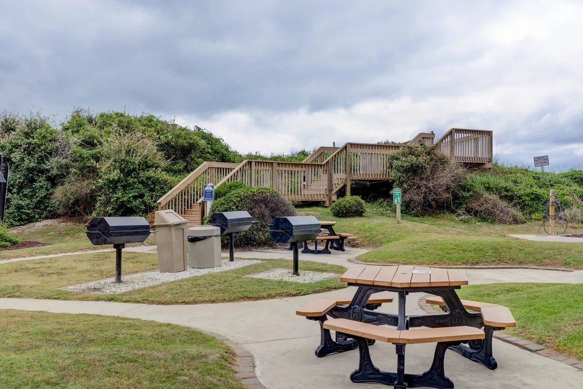 This image captures an inviting outdoor area with picnic tables and barbecue grills, surrounded by greenery. The scene conveys a space designed for family gatherings, meals, and socializing outdoors, with visible amenities for convenient and affordable dining.
