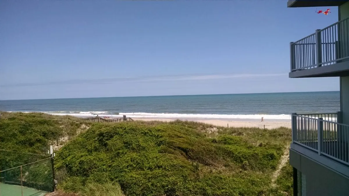 The hero image showcases the condo’s private balcony overlooking sweeping dunes, the sandy shoreline, and the sparkling Atlantic Ocean at North Topsail Beach. The inviting scene highlights easy access to the beach, serene ocean views, and the promise of relaxation and seaside activities right outside the property.