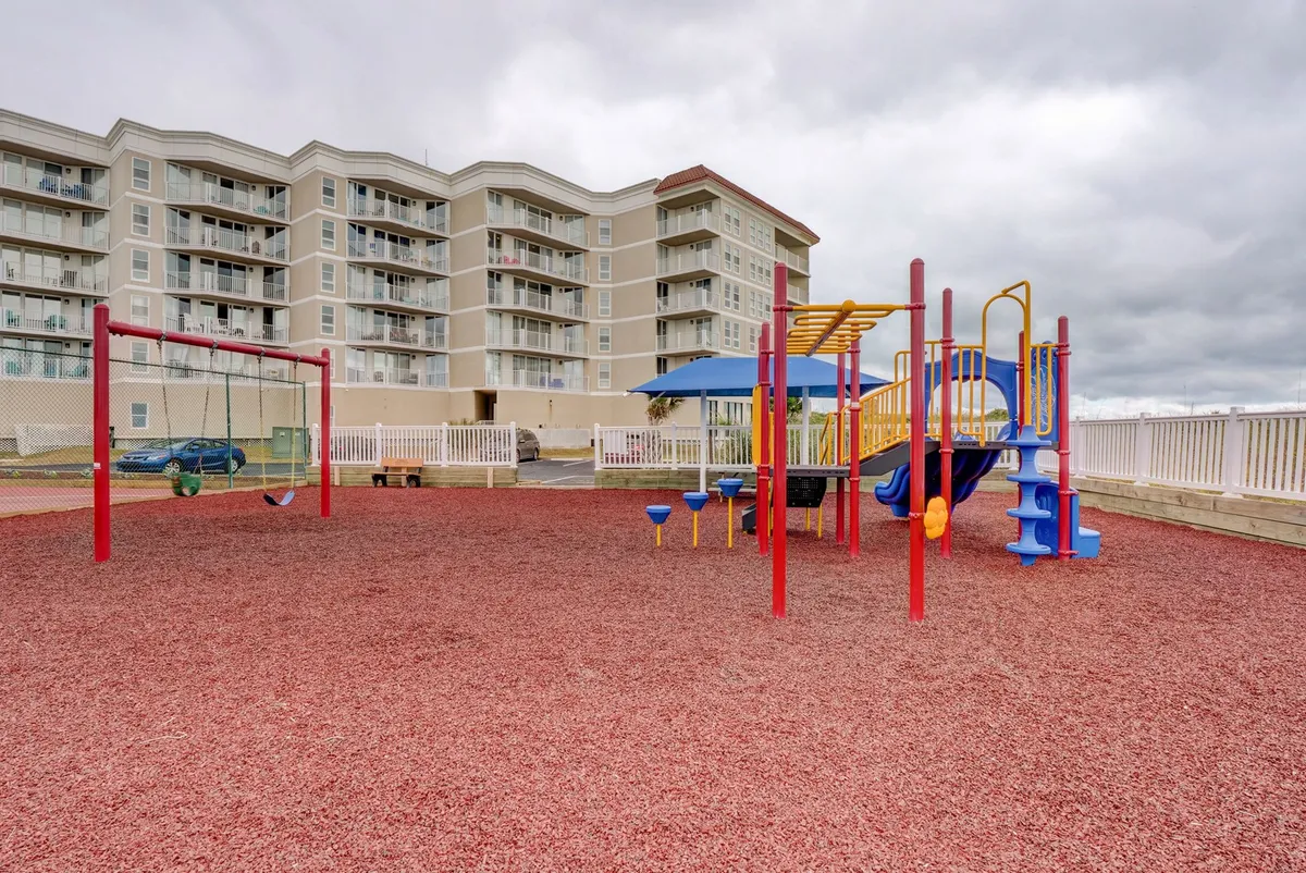 A joyful scene centered on colorful playground equipment and a swing set, set on red safety flooring. Children are shown playing while parents supervise, creating a lively and welcoming space dedicated to family togetherness and imaginative play.
