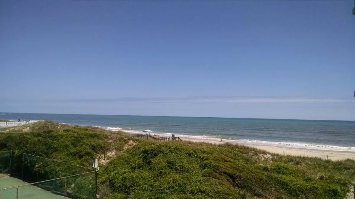 A sweeping view from the property reveals the vast ocean framed by lush green vegetation and a gentle sandy beach stretching toward the horizon. The tranquil scene suggests early morning or sunset, ideal for relaxing while watching the waves or strolling along the shoreline.