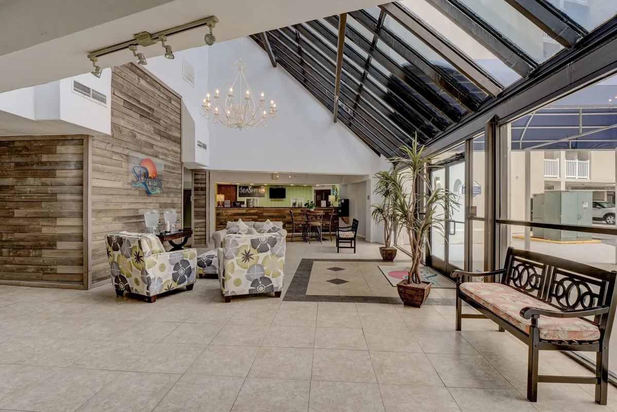 A stylish interior scene featuring a warm, inviting lounge area with a chandelier, plush lounge chairs, a reception desk, and a large potted plant. Natural light pours through expansive windows, creating a bright, comfortable atmosphere that highlights modern coastal decor and sets a tone of relaxation and welcome.