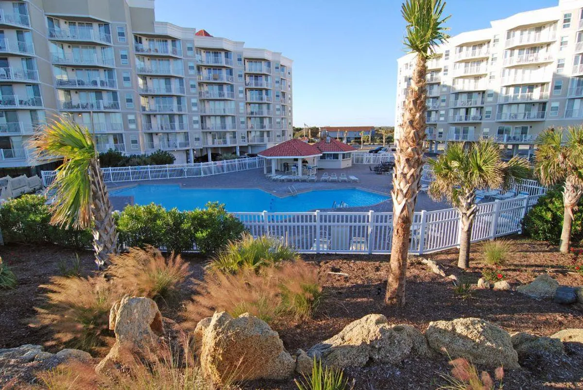 A sparkling swimming pool surrounded by palm trees and landscaped gardens, with a modern apartment building and a stylish cabana topped with a red roof. Lounge chairs are arranged poolside for relaxation, and the atmosphere is inviting and tranquil, suggesting the perfect spot for year-round swimming and socializing.